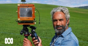 A man with grey hair and a beard smiles beside a vintage camera set on a tripod in a lush green field under a bright sky.