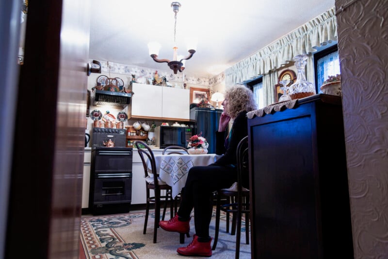 A person with curly hair, dressed in dark clothing and red shoes, sits in a kitchen at a table adorned with a floral centerpiece. The kitchen has a vintage decor, with a patterned carpet, white cabinets, and various trinkets and plates on display.