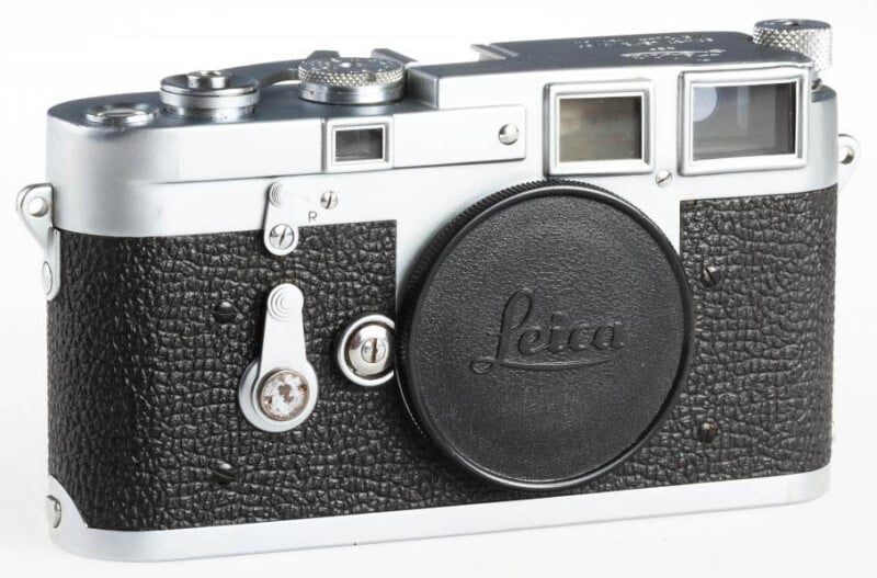 A vintage Leica rangefinder camera with a silver metal top and bottom and a black textured body. The lens cap, bearing the Leica logo, is in place. Various dials and buttons are visible on the top, and there's a viewfinder on the right side.