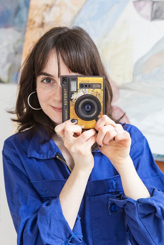 A person with shoulder-length dark hair, wearing a blue shirt and hoop earrings, looks directly at the camera while holding up a vintage-looking camera to their face. The background consists of abstract art pieces.