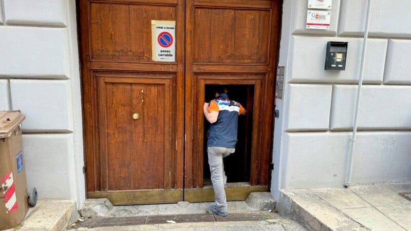 A person in a blue and orange shirt is entering a large wooden door of a building, beside which are a trash bin and a no parking sign.