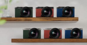 Two wooden shelves each displaying three cameras. The upper shelf holds a green, red, and blue camera, while the lower shelf also holds a green, red, and blue camera. Each camera is marked with the "LUMIX" brand and is equipped with a large lens. A plant is partially visible on the left side.