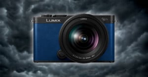 A blue Lumix camera is centered against a backdrop of dark, stormy clouds. The camera lens is prominently displayed, and the brand name "Lumix" is clearly visible above the lens. The overall mood is dramatic, highlighting the camera's sleek design.
