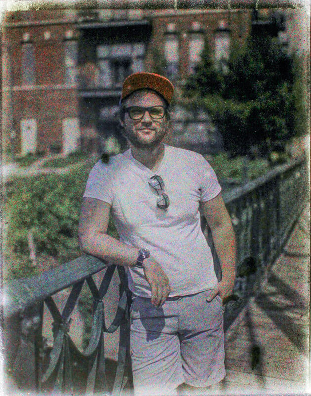 A man in a white t-shirt, sunglasses, and a brown hat smiles while standing by a metal railing with a blurred building in the background. the image has a faded, vintage effect.
