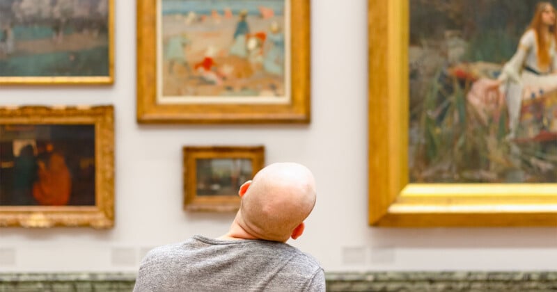 A person stands in front of a gallery wall filled with framed paintings. The individual tilts their head to the side, seemingly observing the artwork closely. The paintings in the background are various sizes and styles.