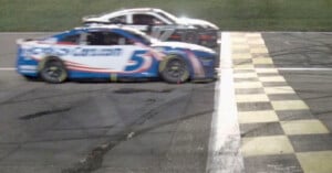 Two race cars speeding past the finish line on a racetrack, with the leading blue and white car crossing over the checkered line.