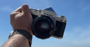 A hand holding a vintage Nikon camera against a clear blue sky. The camera has a large lens and silver and black body. The person is wearing a wristwatch.