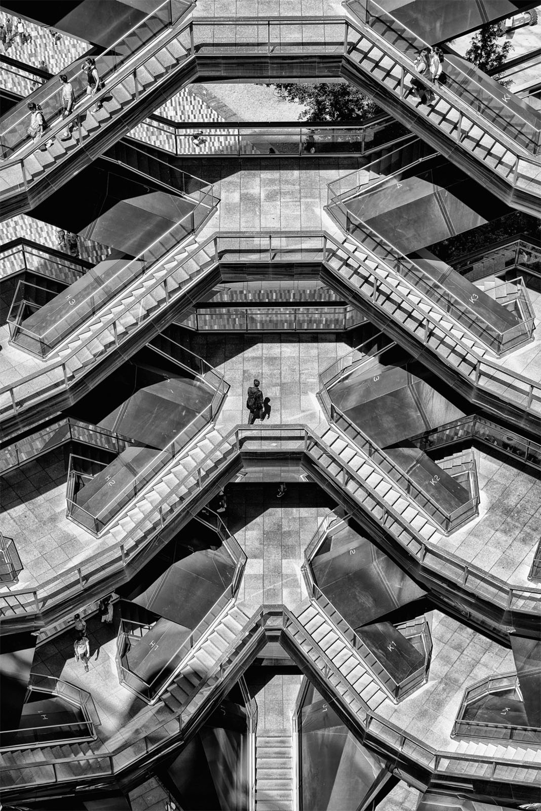 A black and white image of a modern, intricately designed, honeycomb-like structure with multiple levels of walkways and staircases, featuring a few people scattered throughout various levels, emphasizing the geometric pattern and architectural details.