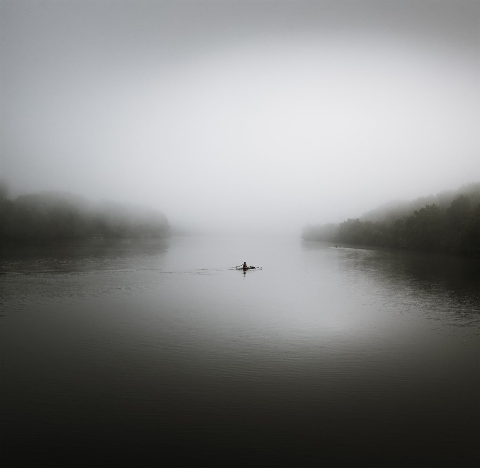 A lone person paddles a small canoe across a calm, misty lake. The dense fog creates a serene and mysterious atmosphere, with barely visible shoreline trees in the distance. The scene is tranquil and moody, evoking a sense of solitude and peacefulness.