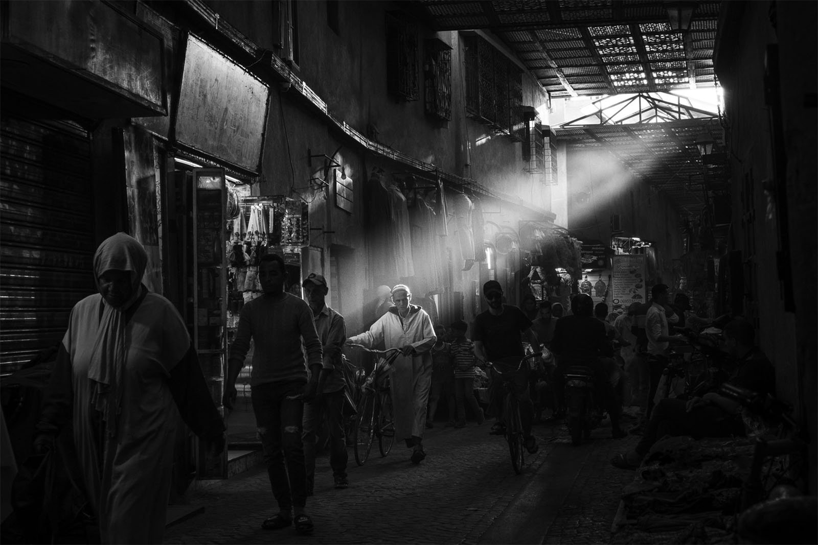 A bustling alley in a marketplace with beams of light streaming through the ceiling. People walk, ride bicycles, and shop at stalls on either side. The scene is in black and white, highlighting the lively yet serene atmosphere of the market.