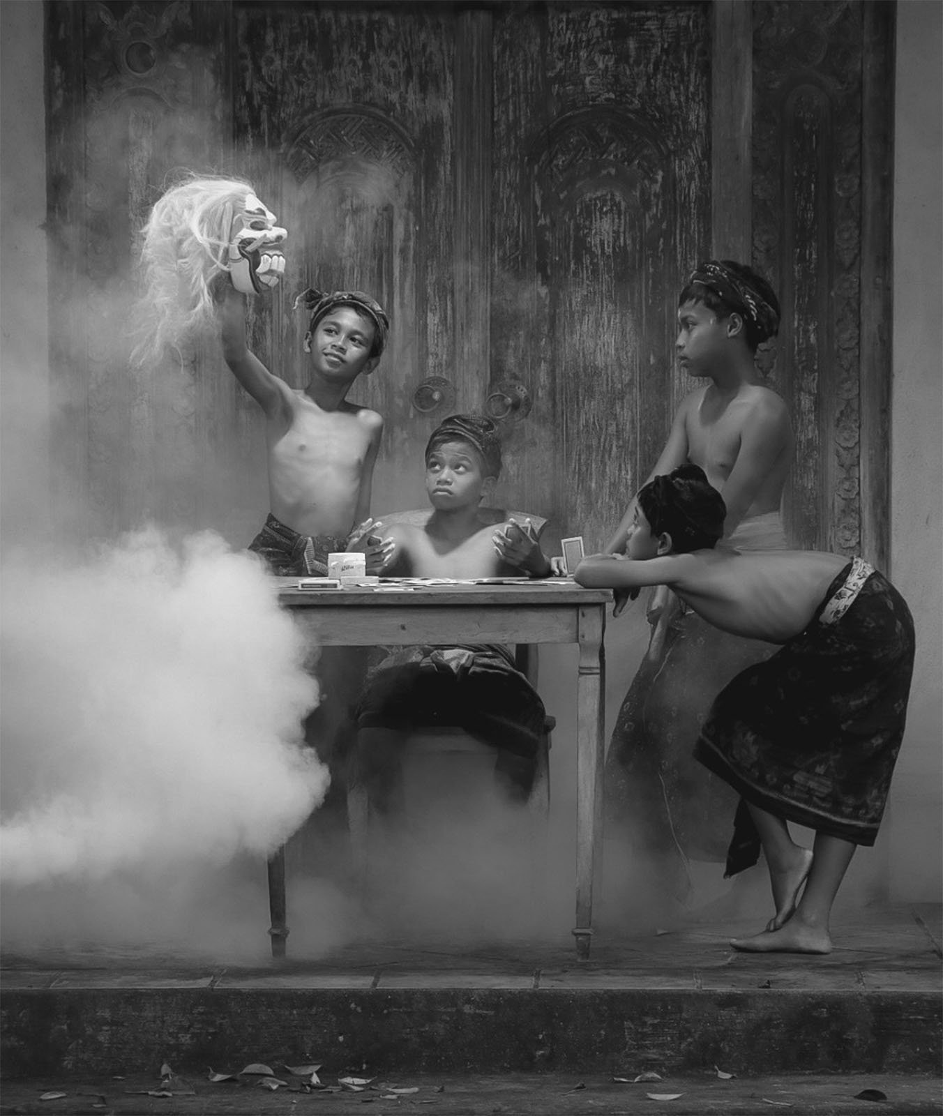 A black-and-white photograph depicts four children wearing traditional clothing. One child holds up a puppet head while others look on with interest. They are gathered around a wooden table, surrounded by a mysterious mist or smoke. The setting appears to be outdoors.