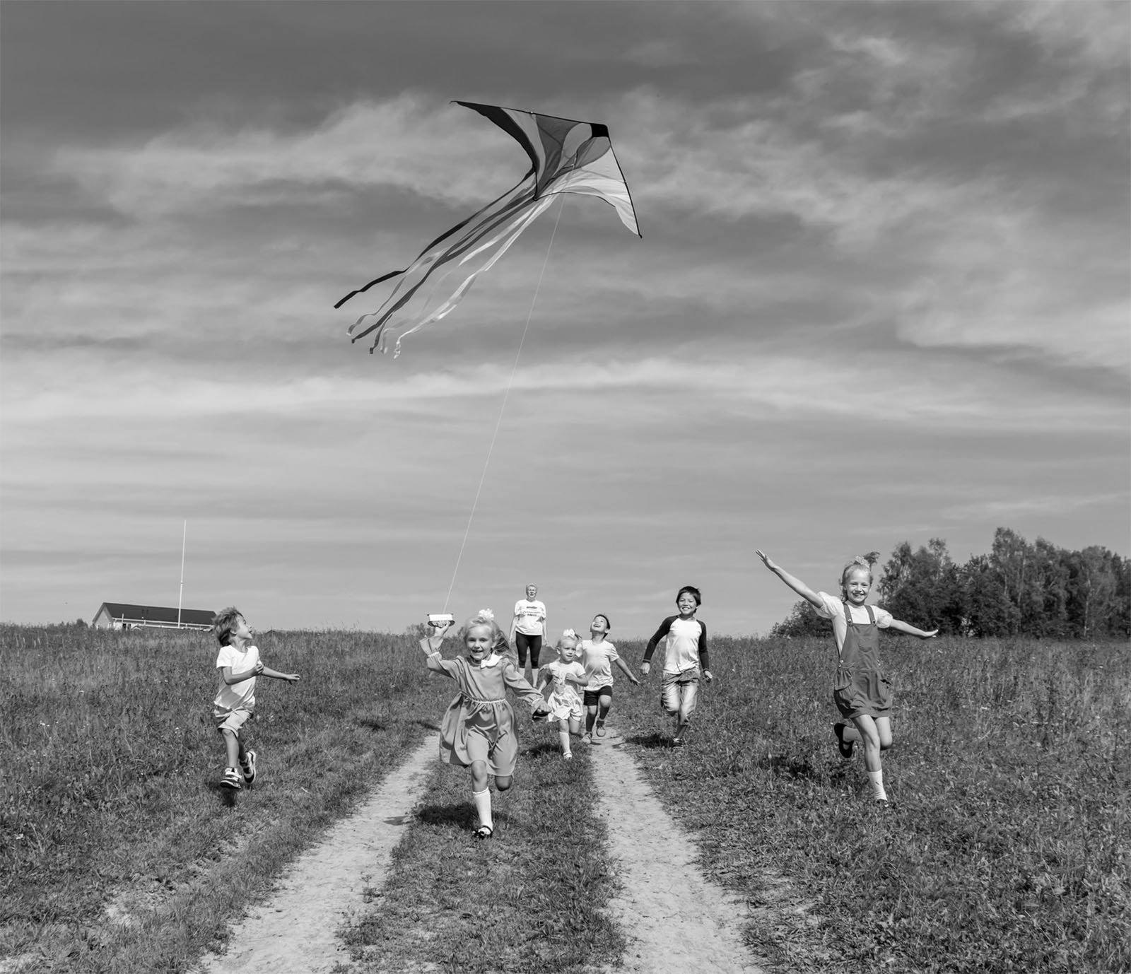 A group of children run excitedly down a dirt path in a grassy field, flying a large kite. The sky above them is partly cloudy, and a small building is visible in the background. The scene is lively and joyful, capturing the fun of outdoor play.