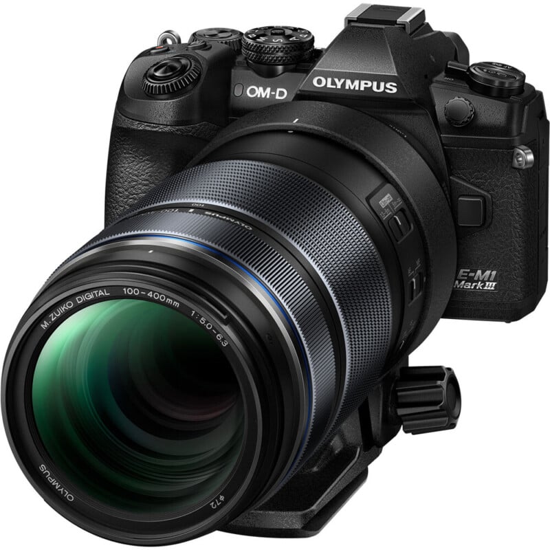 A close-up of a black olympus om-d e-m1 mark iii camera with a m.zuiko 100-400mm telephoto lens attached. the camera features multiple control dials and buttons.