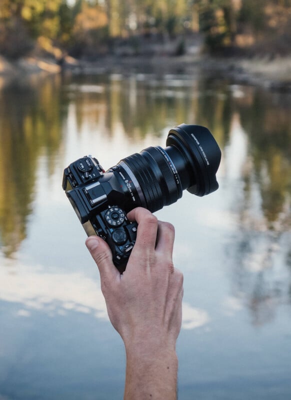 A hand holding a camera with a large lens, extending towards a scenic river surrounded by trees under a clear sky. the camera is in focus, with the lush background softly blurred.
