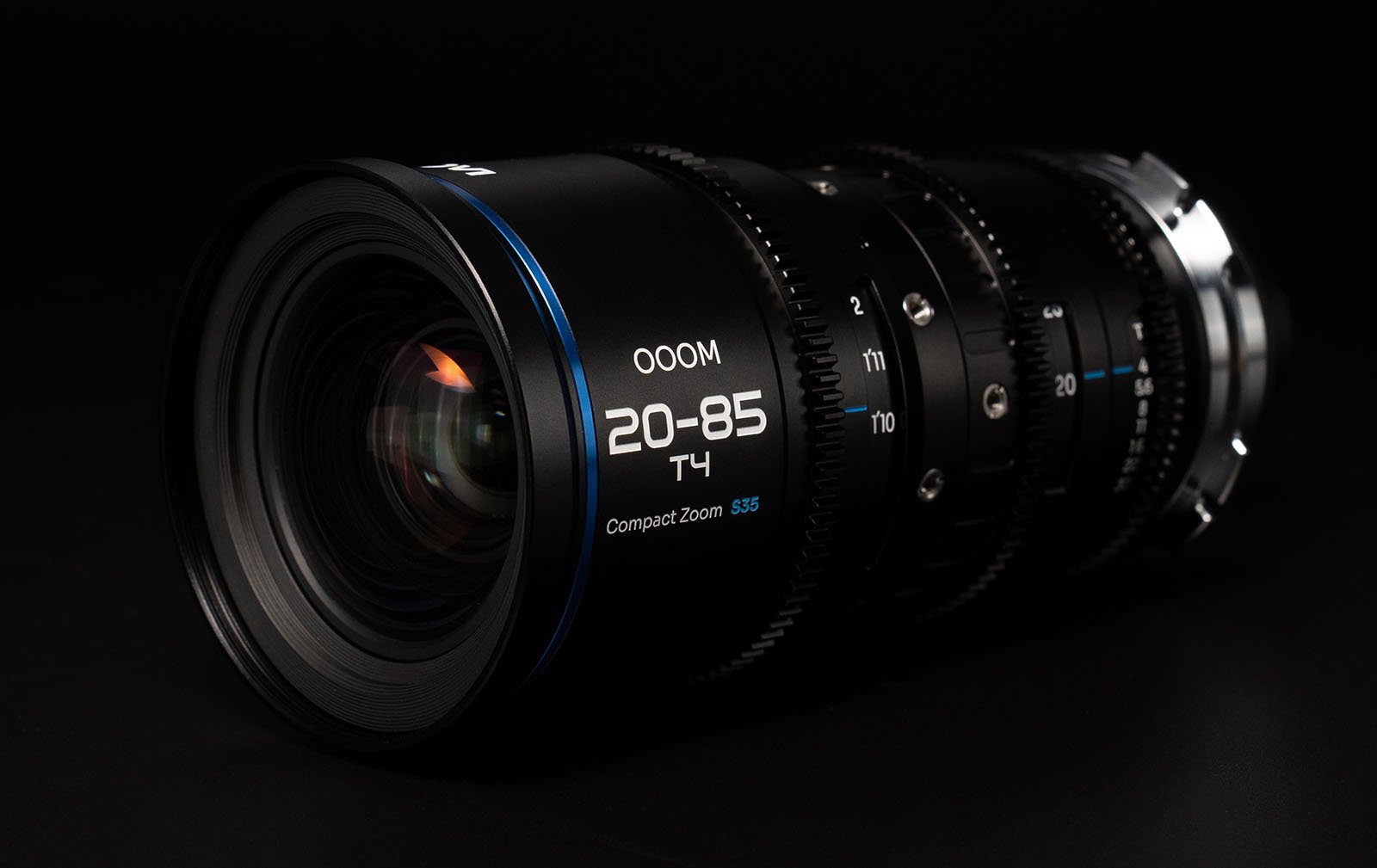 Close-up of a black camera lens labeled "20-85 T4" with various rings for adjusting focus, zoom, and aperture settings. The lens features a reflective front element and blue detailing. The background is black, emphasizing the sleek design of the lens.