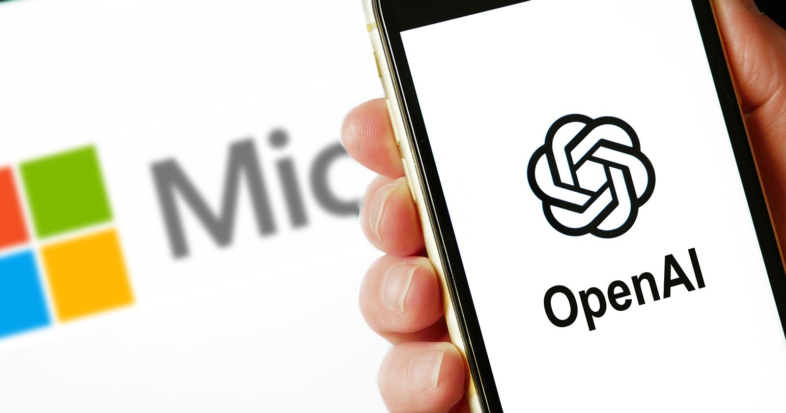 A person holds a smartphone displaying the openai logo, with a blurred microsoft logo visible in the background on another device.