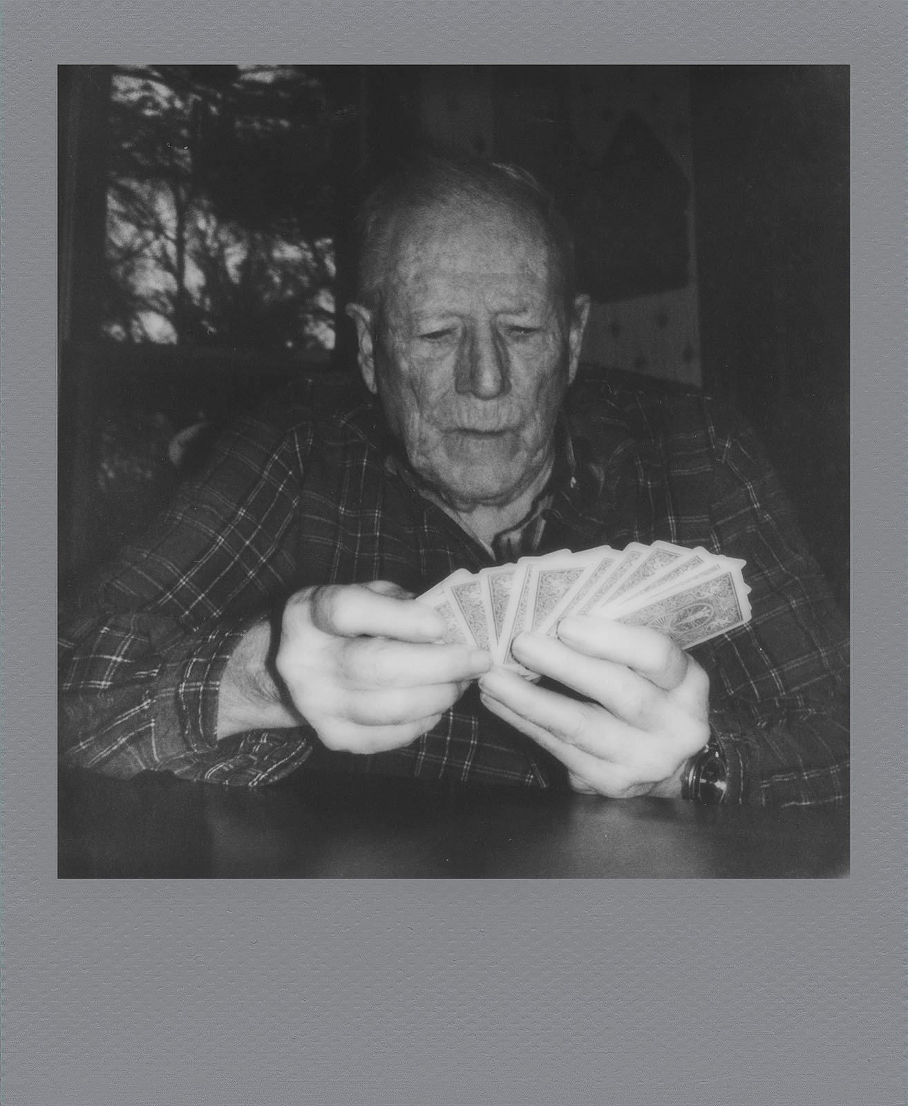 Elderly man sitting at a table, concentrating as he looks at a hand of playing cards. the setting appears indoors, and the photo has a vintage, black and white polaroid style.