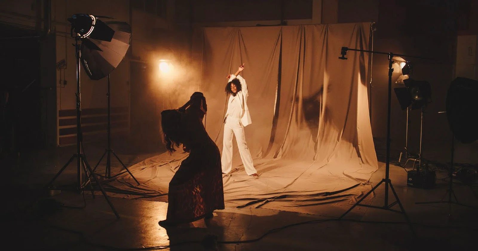 A woman in a white suit poses confidently on a dimly lit set with dramatic fabric drapes behind her. Another person silhouettes against the light, seemingly photographing or observing the scene. Studio lights and equipment surround them, casting warm, moody tones.