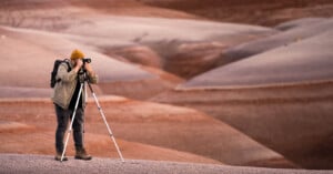 A person, wearing a yellow beanie and backpack, is standing on a rocky terrain, taking photos with a camera mounted on a tripod. The background features a vast, undulating landscape with shades of brown, red, and gray hills.