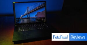 A laptop with an illuminated keyboard is open on a dark surface. The screen displays a vivid image of a bridge over water during twilight. "PetaPixel Reviews" is written in a blue and white banner at the bottom right corner.
.