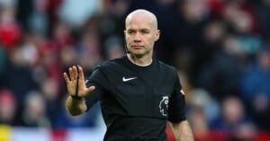 A bald male referee wearing a black uniform with a premier league logo gestures with his right hand on a football field, with blurred spectators in the background.