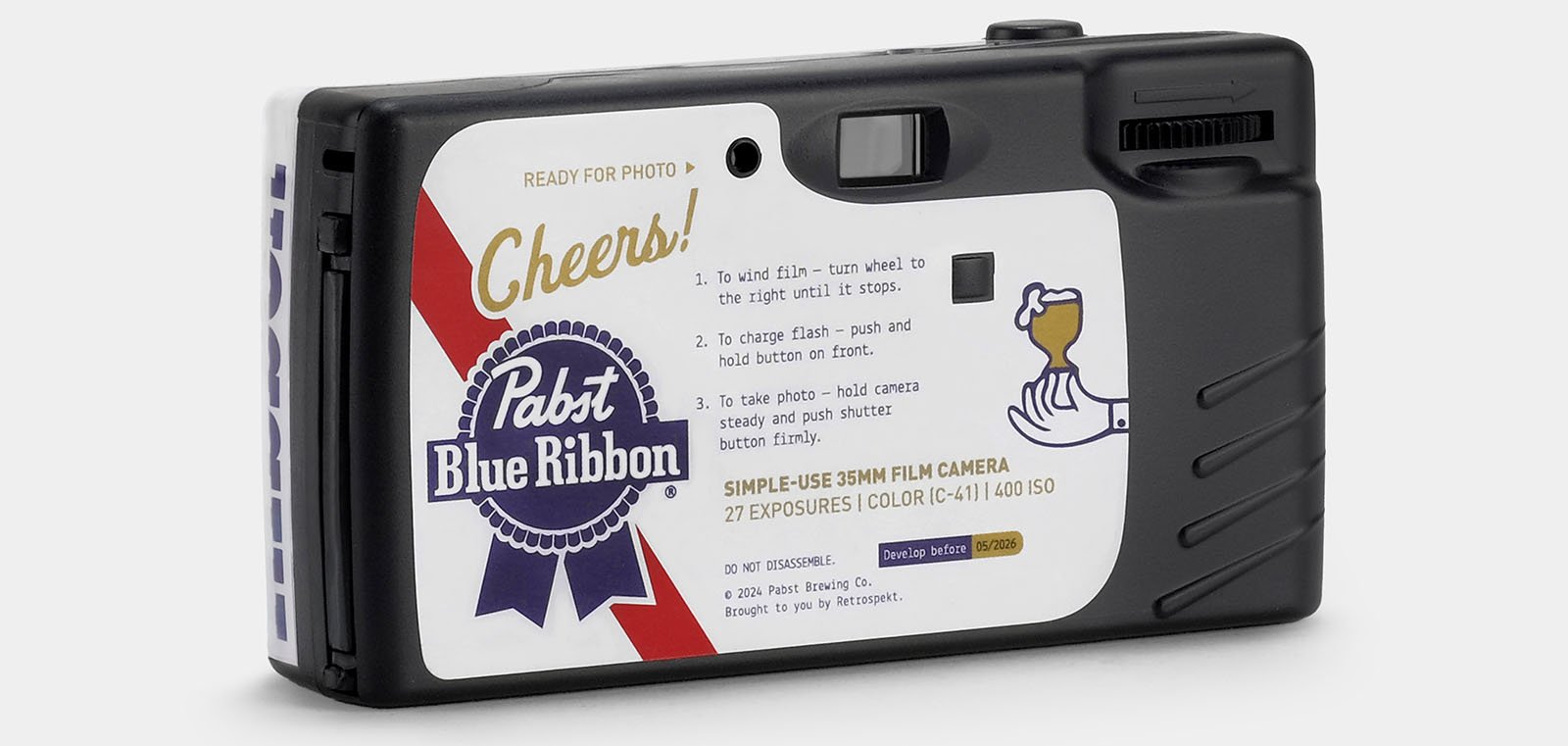 A black disposable 35mm film camera with a white sticker on the back. The sticker features brand elements of Pabst Blue Ribbon, including the logo, a ribbon, and a hand holding a glass. Instructions for use are also printed on the sticker.
