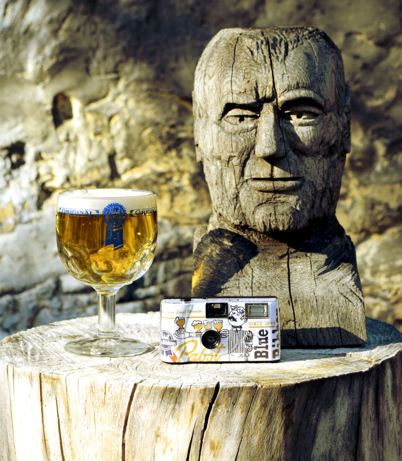 A wooden bust of a man's head, a glass of beer with a frothy top, and a decorated camera are displayed on a cut tree stump against a stone wall.