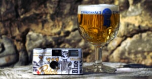 A vintage camera with a Pabst Blue Ribbon design is placed on a surface beside a glass of golden beer with a foamy head. The beer glass is labeled "Original." The background features a stone wall with sunlight casting warm tones on the scene.