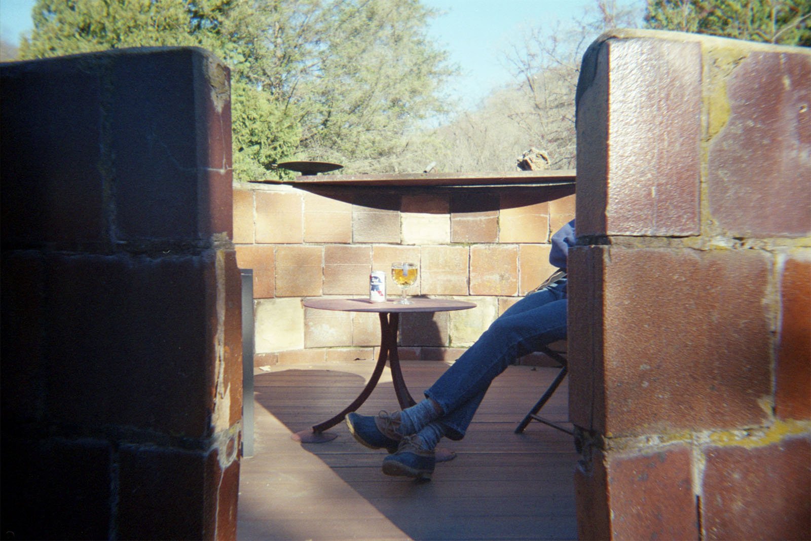 A person sits at an outdoor table inside a circular brick structure. On the round table are a can of beer and a half-full beer glass. The person's legs are crossed, and trees and sky are visible in the background. The scene evokes a relaxed and casual setting.