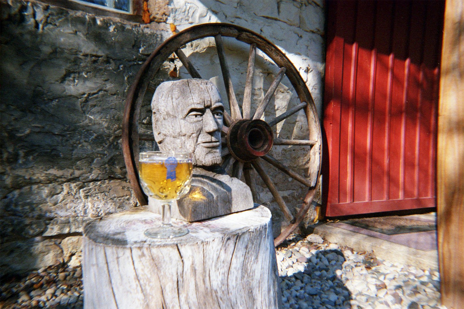 A stone carved head and a wooden wagon wheel are propped against a stone wall. In the foreground, a glass of beer sits atop a cut tree stump. A red door is partially visible on the right side of the image, casting shadows on the scene.