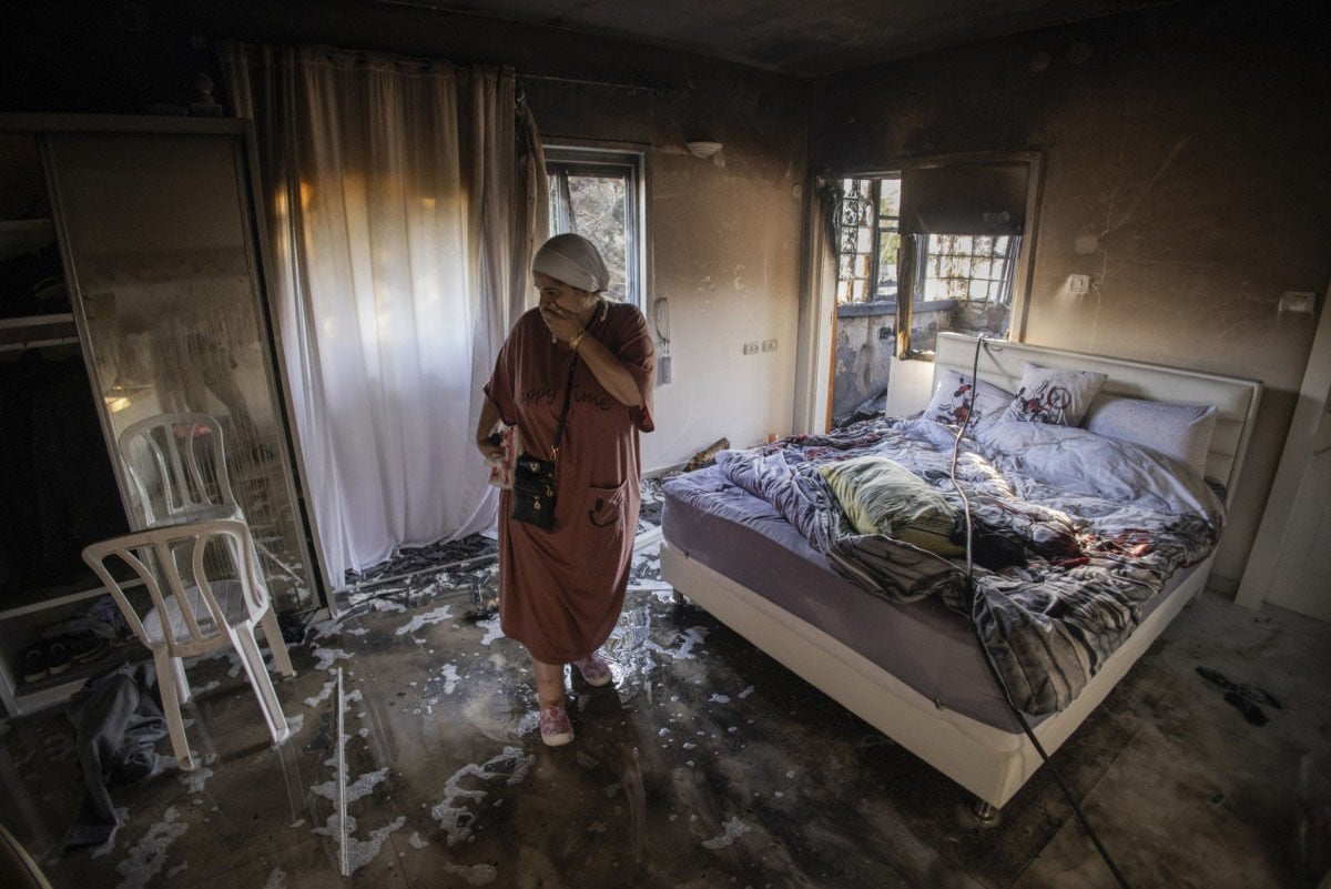 A woman wearing a robe and headscarf stands in a fire-damaged bedroom, holding a camera while looking at charred walls, burnt furniture, and a messy bed with intact curtains.