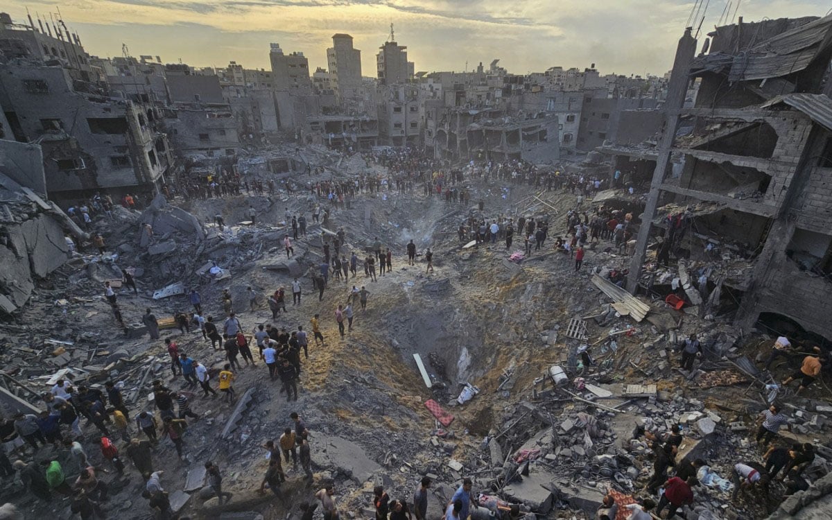Aerial view of a devastated urban area with large crowds gathering around an expansive crater surrounded by demolished buildings under a dusky sky.