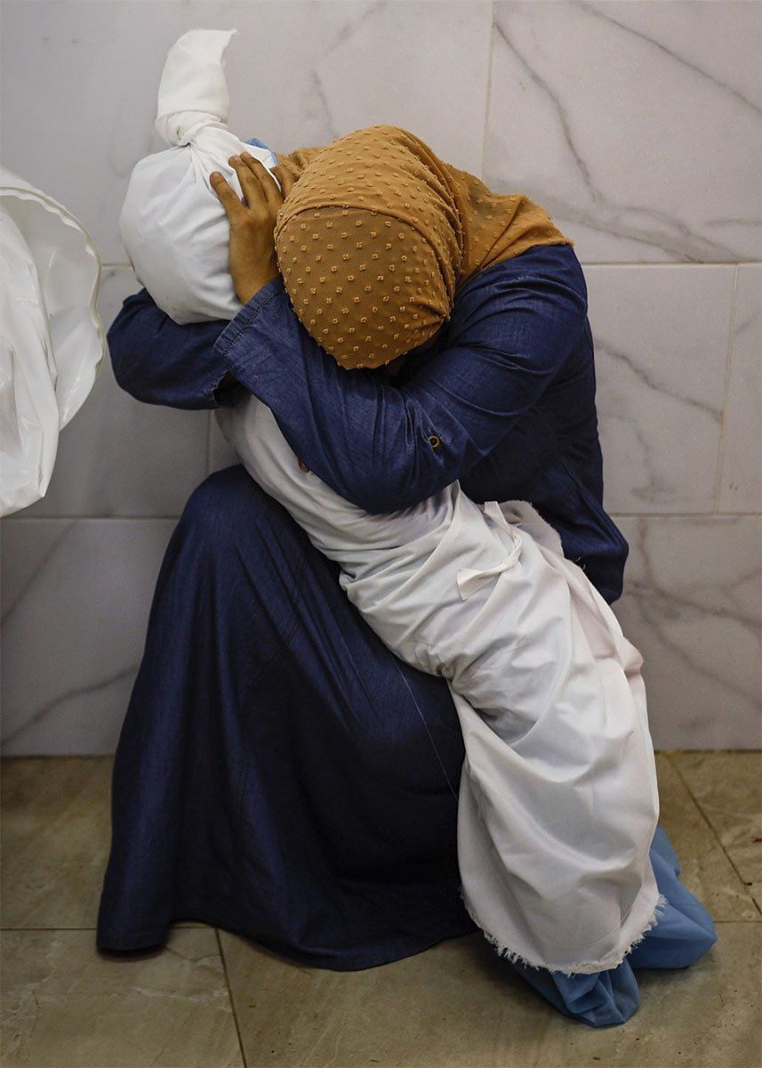 A woman in a headscarf and blue gown cradles a swaddled baby in a hospital, her posture expressing tenderness and care. they sit against a marble wall.