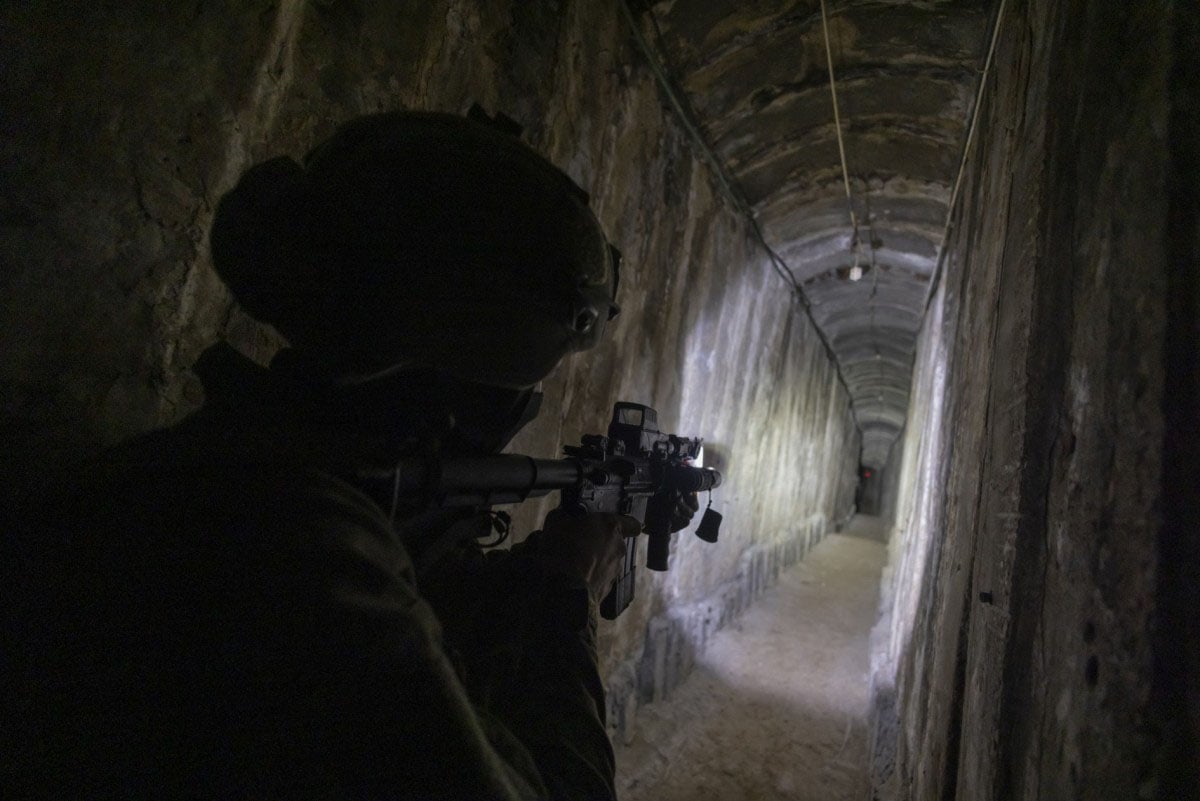 A soldier in tactical gear and helmet aiming a rifle in a dimly lit tunnel, with walls reflecting small amounts of light.