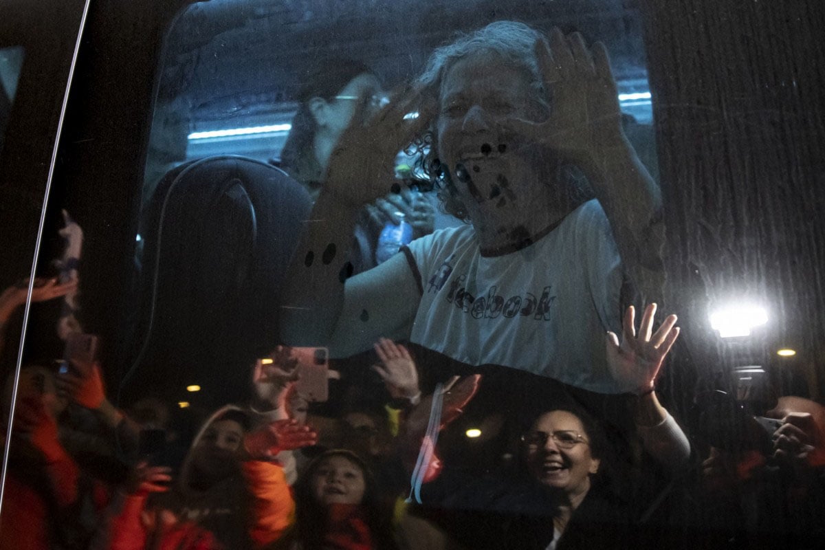A joyful elderly woman presses her hands and face against a window, smiling broadly at people who are observing her from the other side, visible in the reflection.