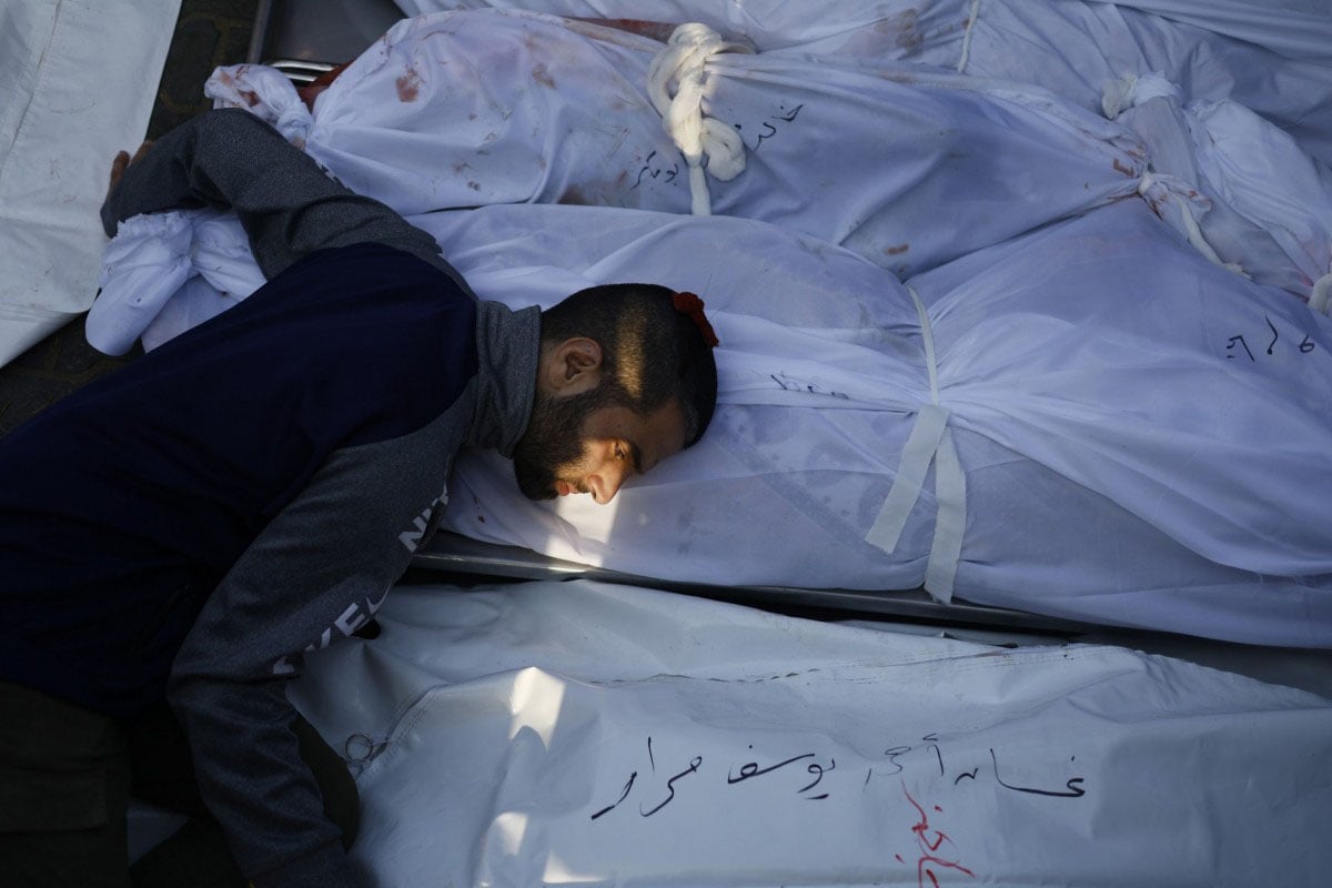 A grieving man rests his head on a shrouded body among several others, all marked with numbers. mourning and loss are palpable in this somber scene.