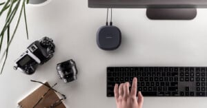 A minimalist workspace showing a person's hand on a black keyboard, a digital camera, lens, portable hard drive, monitor, a notebook, cup, and a plant on a white desk.