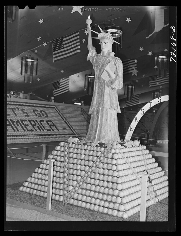 A display at an event features a Statue of Liberty replica made of oranges, set atop a pyramid of oranges. The backdrop displays American flags and the words "Let's go America." Overhead, large stars and additional decorations are visible.