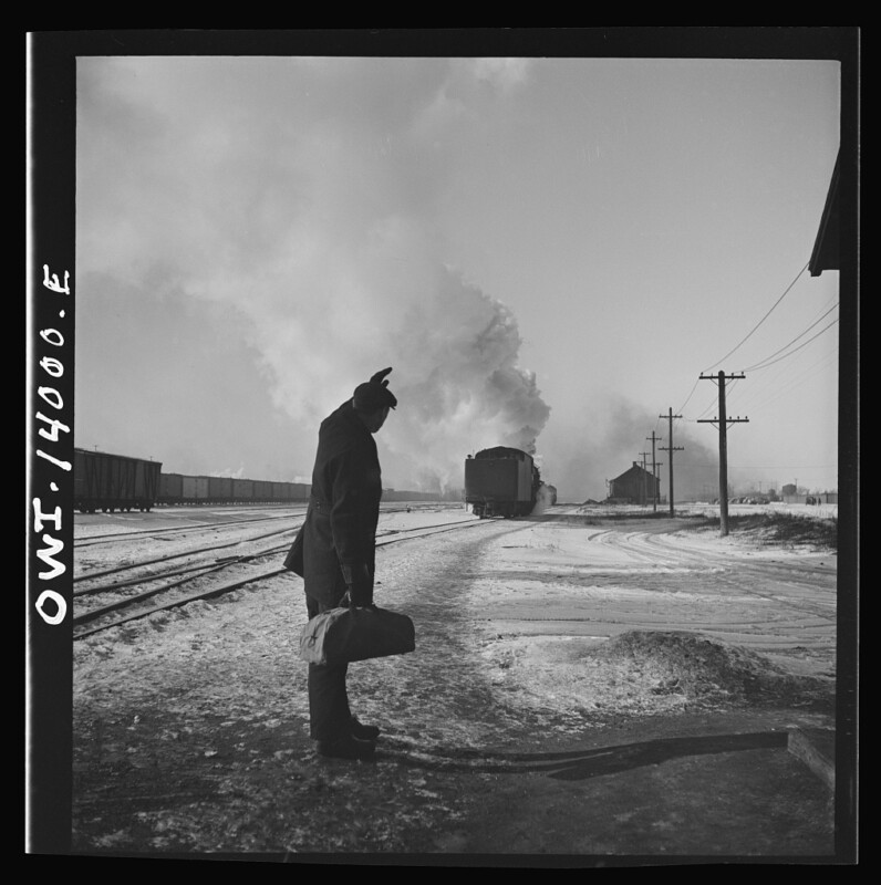 Black and white image of a man standing near snowy railroad tracks with a luggage bag in hand, watching a distant train emitting smoke. Telecommunication poles and a small building can be seen along the tracks in the background.