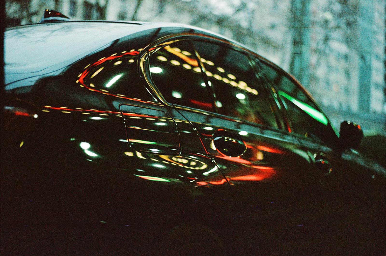 A dark car parked outdoors at dusk with colorful light reflections on its surface, highlighting the sleek curves and glossy finish of the vehicle.
