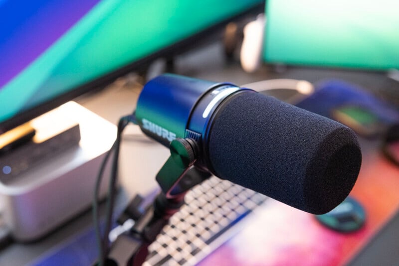 Professional microphone mounted on a stand, positioned in front of a computer keyboard with colorful backlit keys and multiple monitor screens displaying vibrant colors.