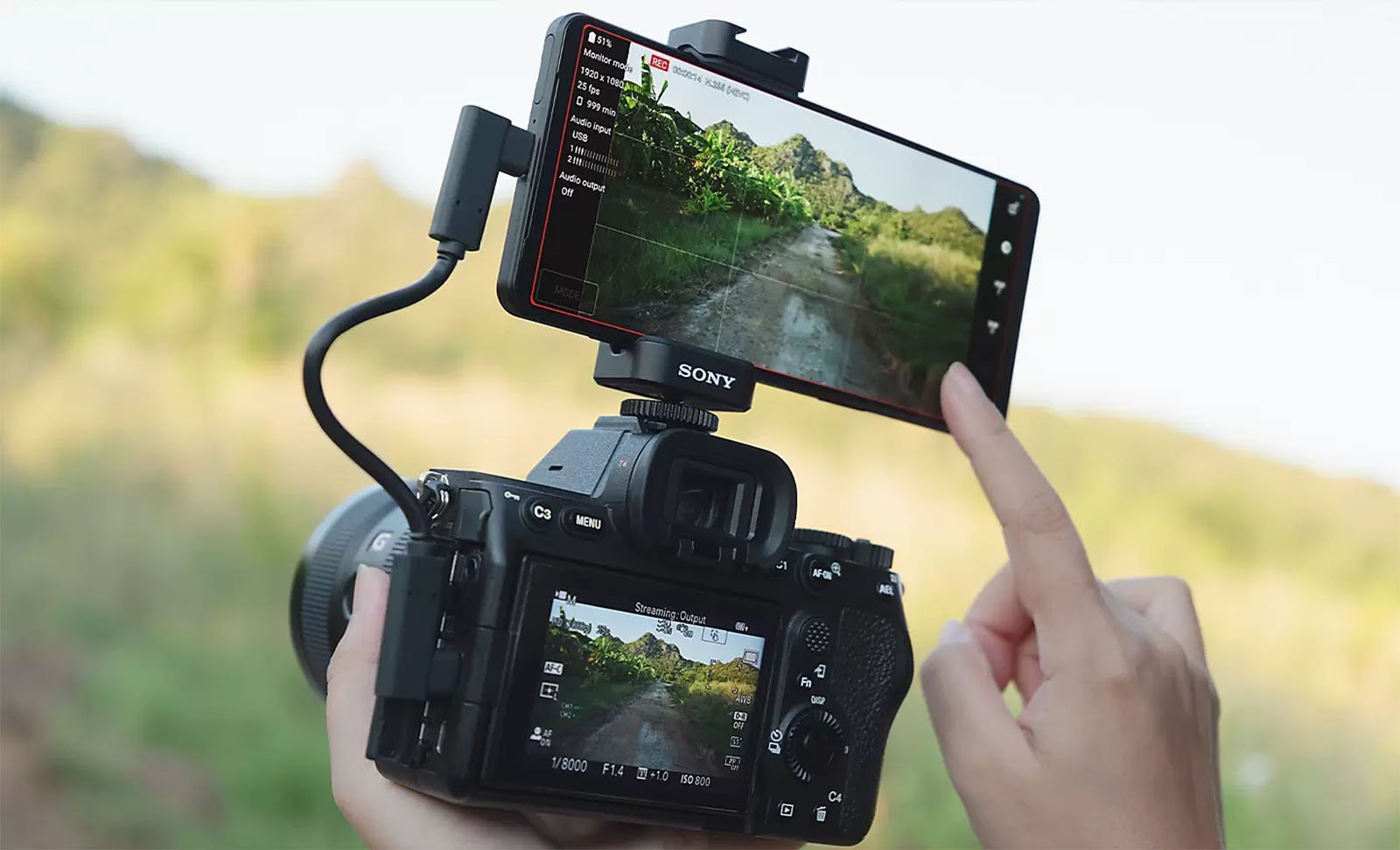 A close-up of a person using a professional camera with an attached monitor displaying the same image as the camera's screen. The monitor shows a scenic outdoor path surrounded by greenery. The camera is branded "Sony," and the person's hand is touching the monitor's screen.