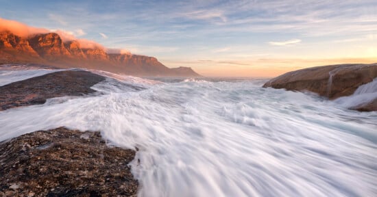 A dynamic seascape showing white waves crashing against dark rocky shorelines. In the background, mountains are bathed in the warm glow of a setting sun under a partly cloudy sky. The scene evokes a sense of power and serenity.