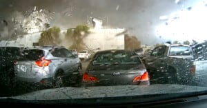View from inside a car showing a tornado take apart a building with a heavy downpour on the windshield with other stationary cars and a building in a parking lot. Water droplets and streaks are visible on the glass.