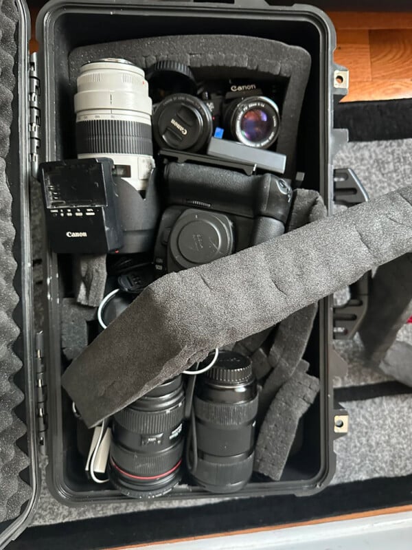 A hard protective case with foam padding holds a collection of camera equipment, including several Canon lenses, a camera body, a flash unit, and various camera accessories. The gear is securely nestled within the foam compartments for safekeeping.