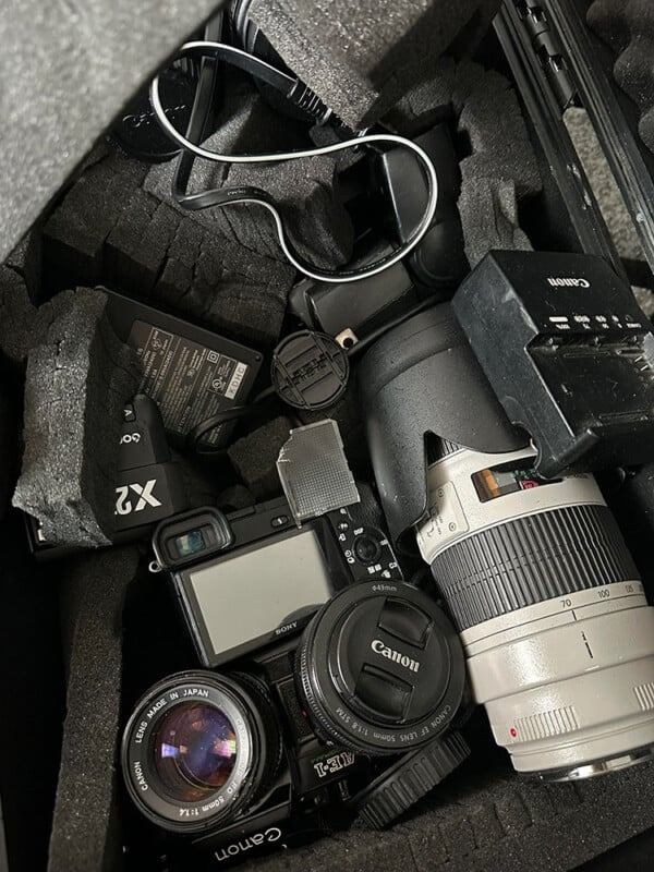 A camera bag with foam padding holding various photography equipment including two cameras, multiple camera lenses, charger, cables, and accessories. The gear appears slightly disorganized within the compartmentalized sections of the bag.