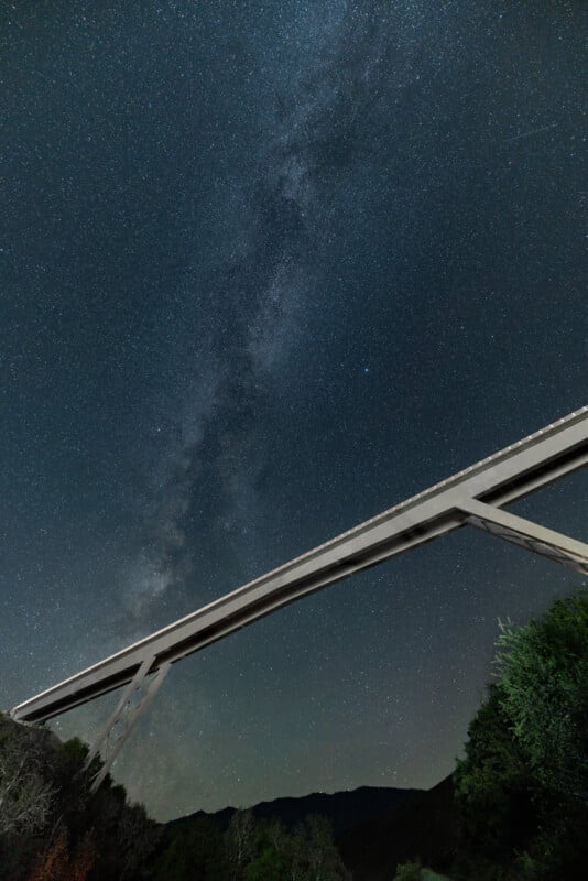 A night sky filled with shimmering stars and the milky way, viewed from beneath a modern bridge extending diagonally across the frame, surrounded by dark silhouettes of trees.
