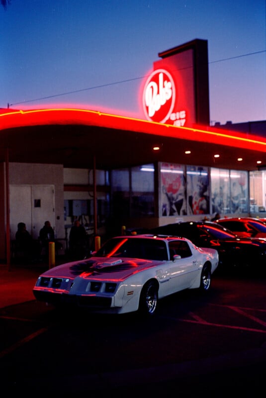 A classic white Muscle Car is parked in front of a retro-styled diner with a brightly lit neon sign that reads "Bob's Big Boy." It's dusk, and the sky has a gradient of blue and purple hues. Other cars are visible in the lot, and the diner has several patrons.