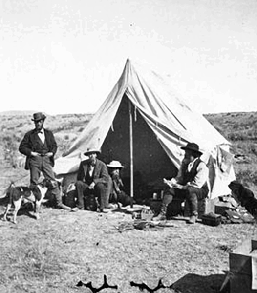 Black and white historical photo of three men and two dogs at a campsite with a large tent in a grassy field. they appear to be in conversation, sitting and standing around scattered camping gear.