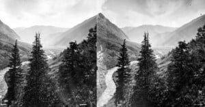 Stereoscopic view of a lush mountain valley with a river running through it and coniferous trees in the foreground; rugged mountain peaks rise in the background.